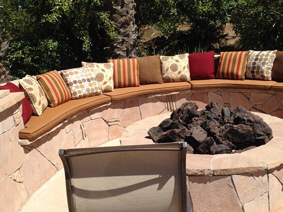 Custom-made curved outdoor bench cushions in tan with decorative patterned toss pillows around a stone fire pit.