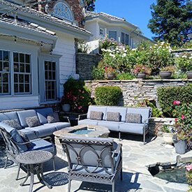 Slate blue outdoor sofa cushions with grey patterned lumbar accent pillows on a metal patio furniture set around a stone fire pit in Los Angeles.